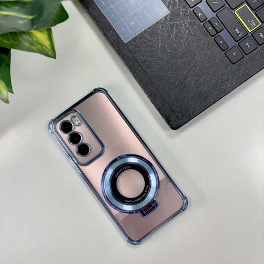 A transparent Oppo Reno 12 pro phone case with a magnetic metal kickstand and blue accents around the ring, placed on a desk next to a laptop and a plant.